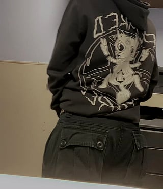In the work restroom, a person wearing a black hoodie with a cartoon character on it is seen standing with their back to the camera.'